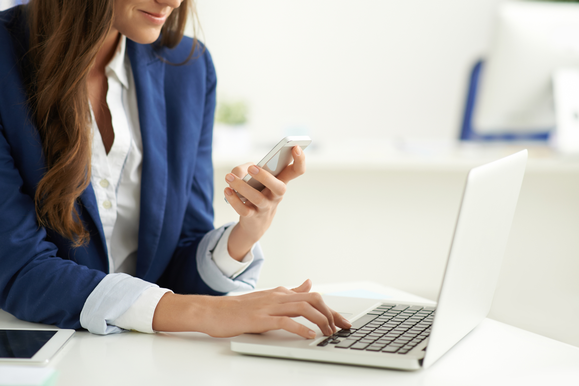 Frau im blauen Blazer arbeitet am Laptop und schaut dabei auf ihr Mobiltelefon.